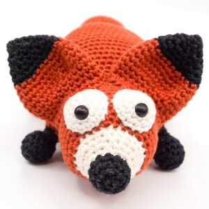 May include: A crocheted fox toy with orange fur, black ears and nose, and white eyes. The toy has a friendly expression.