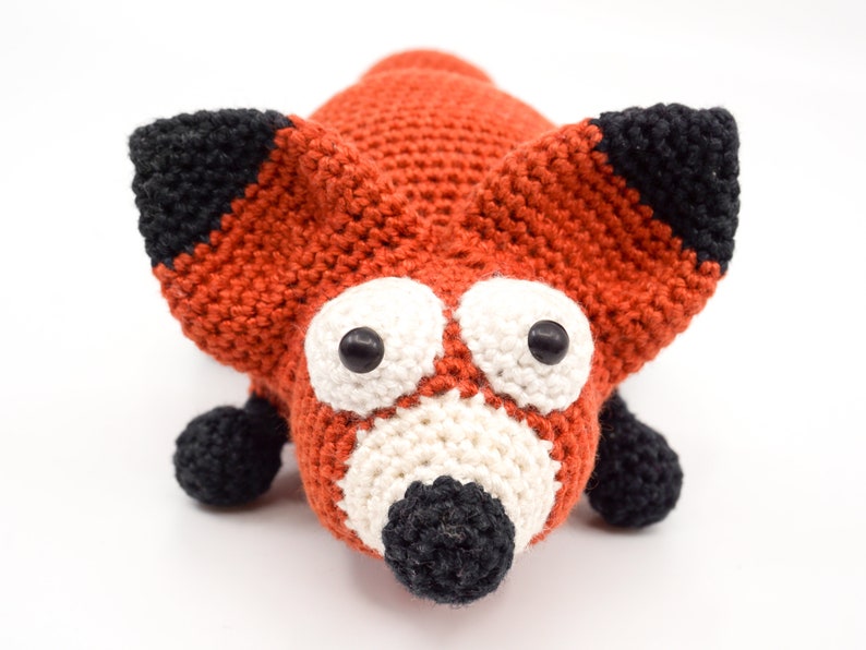 May include: A crocheted fox toy with orange fur, black ears and nose, and white eyes. The toy has a friendly expression.