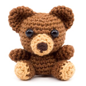 May include: A brown crocheted teddy bear with black eyes and a tan nose. The bear is sitting with its legs crossed and its arms outstretched.