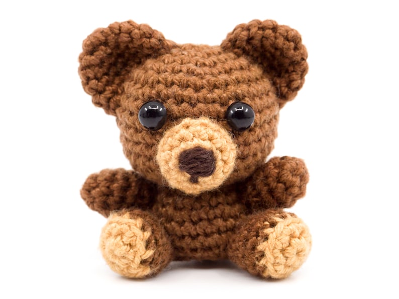 May include: A brown crocheted teddy bear with black eyes and a tan nose. The bear is sitting with its legs crossed and its arms outstretched.
