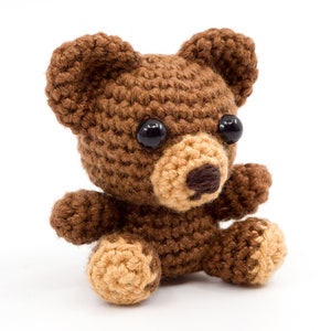 May include: A small brown crocheted teddy bear with black eyes and a tan nose. The bear is sitting on a white surface.