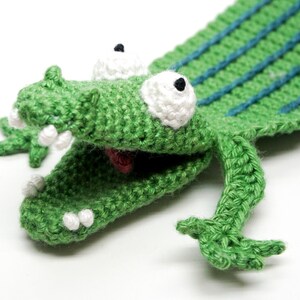 Pu&ograve; includere: Marionetta di coccodrillo in crochet verde con occhi e denti bianchi. La bocca del coccodrilo &egrave; aperta e la marionetta ha una lunga coda.