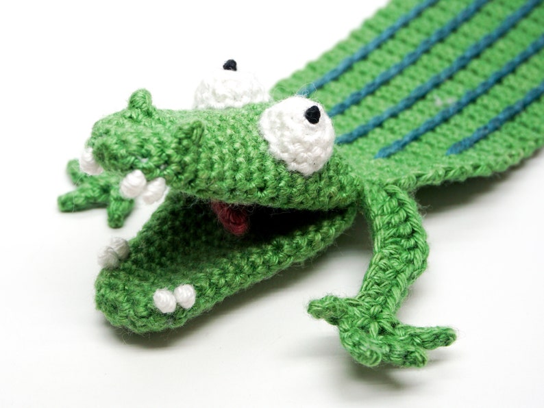 Pu&ograve; includere: Marionetta di coccodrillo in crochet verde con occhi e denti bianchi. La bocca del coccodrilo &egrave; aperta e la marionetta ha una lunga coda.