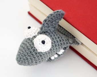 May include: A gray crocheted shark bookmark with white eyes and teeth, peeking out from behind a red and cream book.