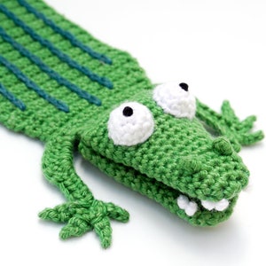 Puede incluir: Marioneta de cocodrilo verde de crochet con ojos blancos y pupilas negras. El cocodrilo tiene una cola larga y una boca abierta con dientes blancos.