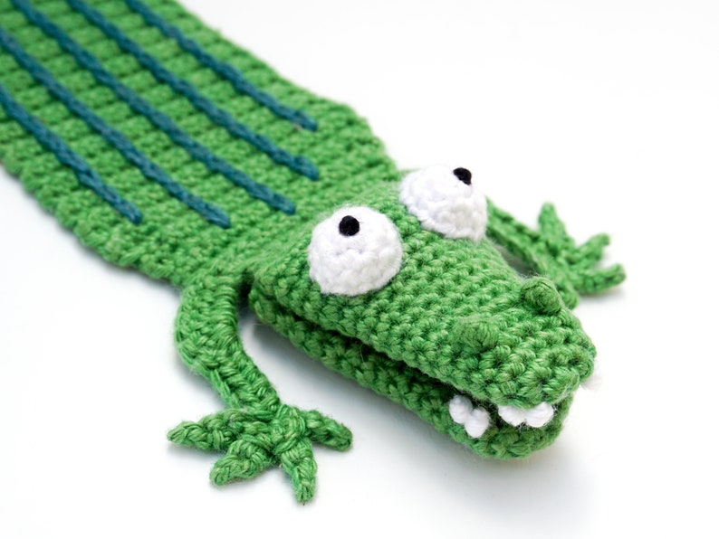 Puede incluir: Marioneta de cocodrilo verde de crochet con ojos blancos y pupilas negras. El cocodrilo tiene una cola larga y una boca abierta con dientes blancos.