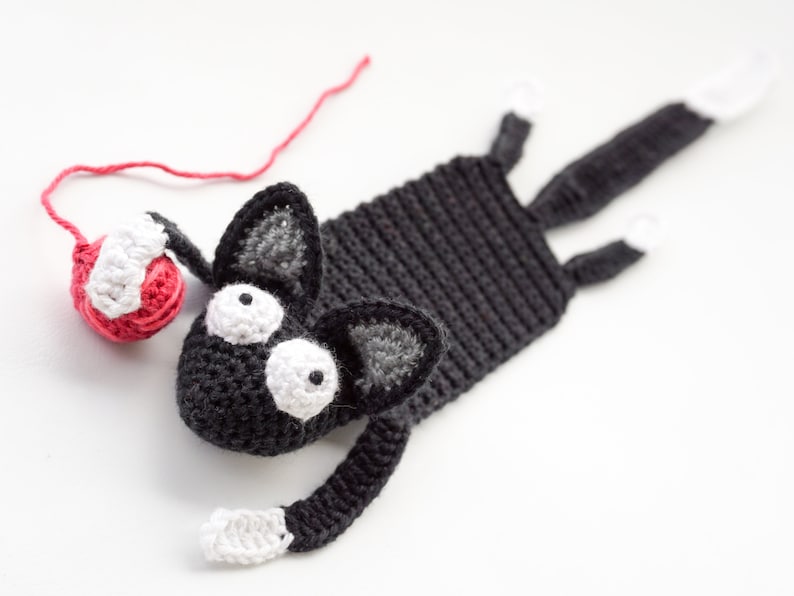 May include: A black crocheted cat bookmark with white yarn accents and large white eyes. The cat is holding a red yarn ball.