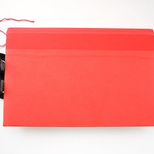 May include: A white crocheted bookmark with black eyes and red yarn peeking out from behind a red folder.