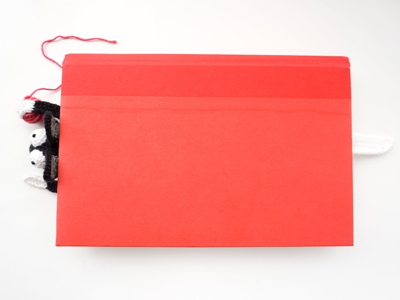May include: A white crocheted bookmark with black eyes and red yarn peeking out from behind a red folder.
