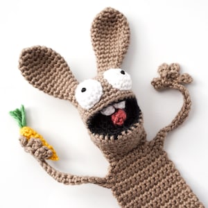 May include: A brown crocheted bunny with large white eyes and a red tongue holds a yellow and green crocheted carrot.