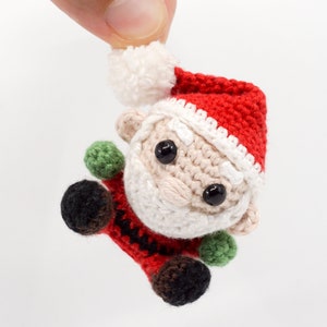 Puede incluir: Un peque&ntilde;o adorno de Pap&aacute; Noel de crochet con un gorro rojo, ribete blanco y barba blanca. El adorno tiene ojos negros, nariz marr&oacute;n y guantes verdes.