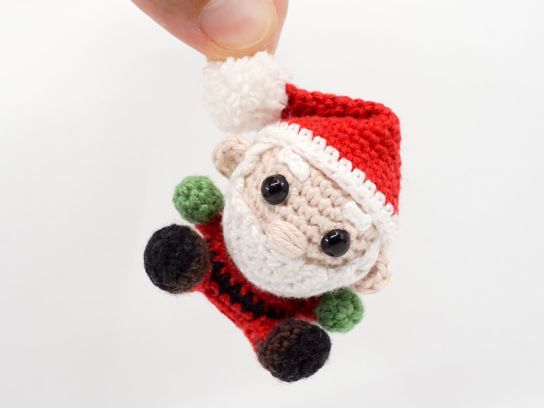 Puede incluir: Un peque&ntilde;o adorno de Pap&aacute; Noel de crochet con un gorro rojo, ribete blanco y barba blanca. El adorno tiene ojos negros, nariz marr&oacute;n y guantes verdes.