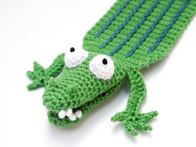 Puede incluir: Marcap&aacute;ginas de cocodrilo verde tejido a crochet con ojos y dientes blancos. El cocodrilo tiene una espalda rayada verde y azul.