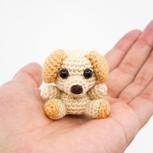 May include: A small crocheted dog toy with brown ears and black eyes. The dog is sitting in the palm of a hand.