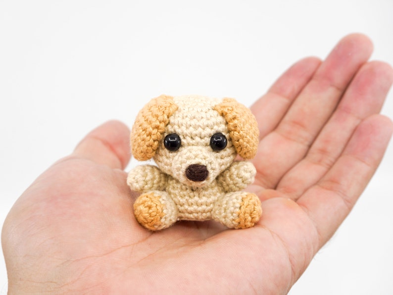 May include: A small crocheted dog toy with brown ears and black eyes. The dog is sitting in the palm of a hand.