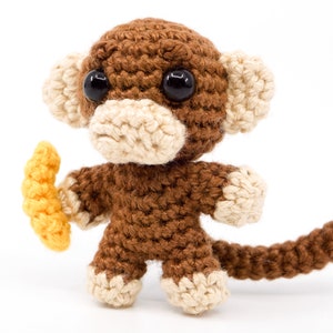 May include: A brown crocheted monkey toy holding a yellow crocheted banana. The monkey has black eyes and a white crocheted nose and mouth.