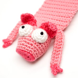 May include: Pink crocheted pig bookmark with large white eyes and black pupils. The pig has a long body and small pink ears.