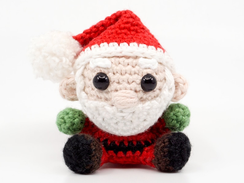 Puede incluir: Un adorno de Pap&aacute; Noel de crochet con un gorro rojo, barba blanca y ojos negros. El adorno est&aacute; sentado sobre una superficie blanca.