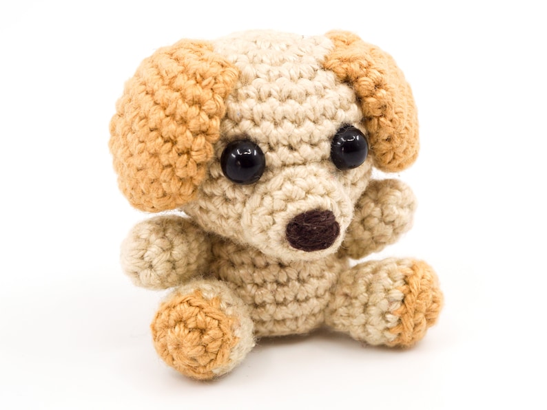 May include: A small crocheted dog toy with brown eyes, a brown nose, and tan ears. The toy is sitting on a white background.