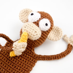Puede incluir: Un marcapáginas de mono de crochet marrón que sostiene un plátano amarillo. El mono tiene ojos blancos y una nariz marrón.