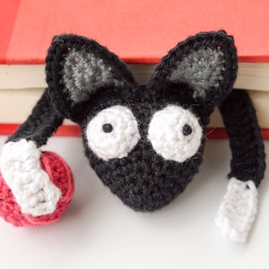 May include: A black and white crocheted cat bookmark with large white eyes and a red yarn ball. The bookmark is peeking over the edge of a red book.