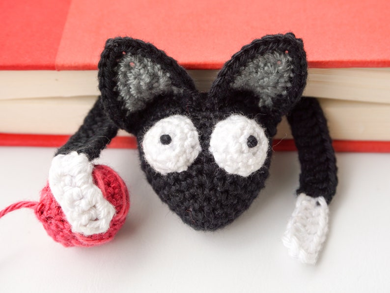 May include: A black and white crocheted cat bookmark with large white eyes and a red yarn ball. The bookmark is peeking over the edge of a red book.