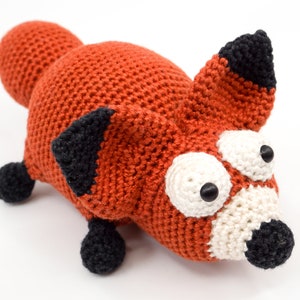 May include: A crocheted fox plush toy with large, white eyes and a black nose. The fox is orange with black ears and feet.