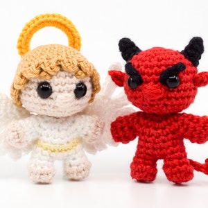 May include: A white crocheted angel with a yellow halo and a red crocheted devil with black horns. Both figures have black eyes and are standing on a white surface.