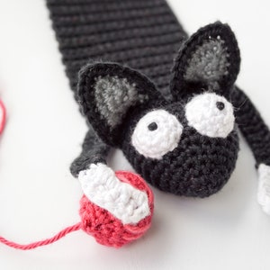 May include: A black crocheted cat bookmark holding a red yarn ball. The cat has white eyes with black pupils and white paws.