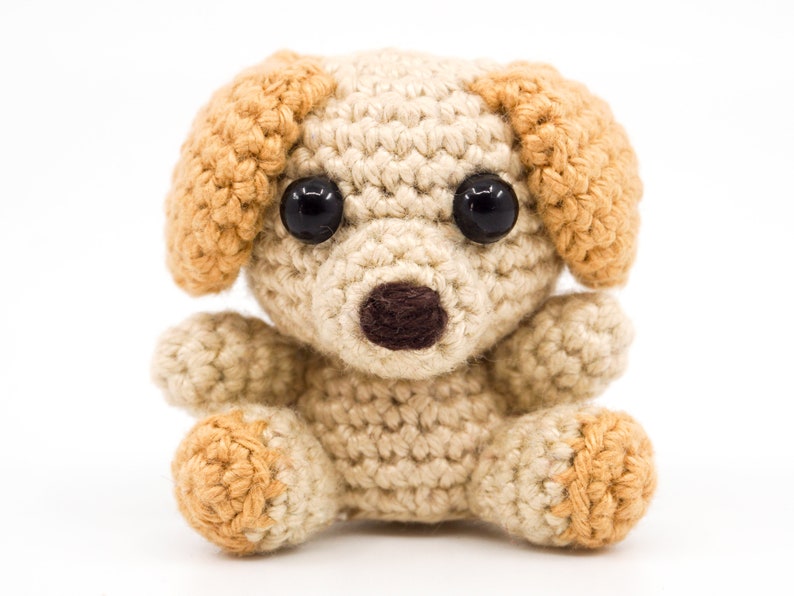 May include: A small crocheted dog toy with brown eyes, a brown nose, and tan ears. The dog is sitting on a white background.
