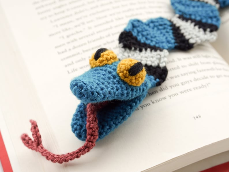 Pu&ograve; includere: Un segnalibro a forma di serpente all'uncinetto blu con strisce bianche e nere, occhi gialli e lingua rossa. Il segnalibro &egrave; appoggiato su un libro.