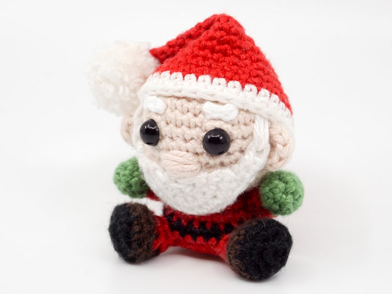 Puede incluir: Un mu&ntilde;eco de Pap&aacute; Noel de crochet con un gorro rojo, barba blanca y ojos negros. El mu&ntilde;eco lleva un traje rojo y blanco con detalles verdes.