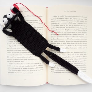 May include: A black crocheted cat bookmark with white crocheted paws and a red yarn ball. The bookmark is placed on an open book with text about weddings.