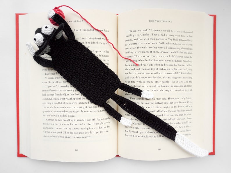 May include: A black crocheted cat bookmark with white crocheted paws and a red yarn ball. The bookmark is placed on an open book with text about weddings.