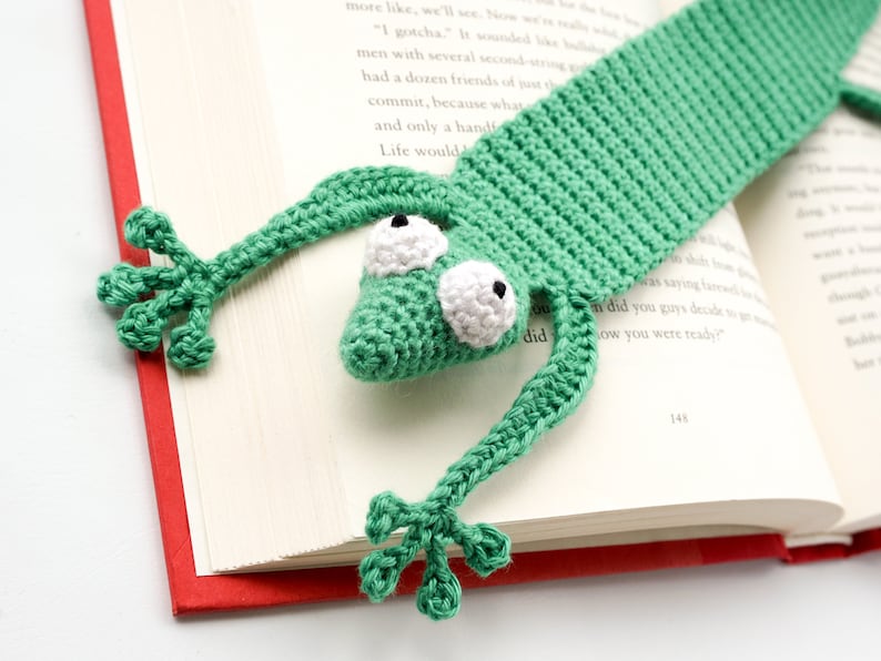 Pu&ograve; includere: Segnalibro a forma di rana lavorato a uncinetto verde con occhi bianchi. Il segnalibro &egrave; appoggiato su un libro chiuso con una copertina rossa.