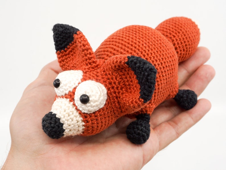 May include: A crocheted fox toy with large white eyes and black accents. The fox is orange and has a white nose and mouth.
