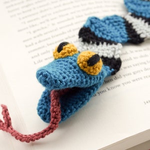 May include: A blue crocheted snake bookmark with black and white stripes, yellow eyes, and a red tongue. The bookmark is lying on a book.