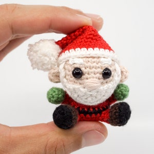 Puede incluir: Un peque&ntilde;o adorno de Pap&aacute; Noel de ganchillo con un gorro rojo, barba blanca y ojos negros. El adorno est&aacute; sentado en una mano.