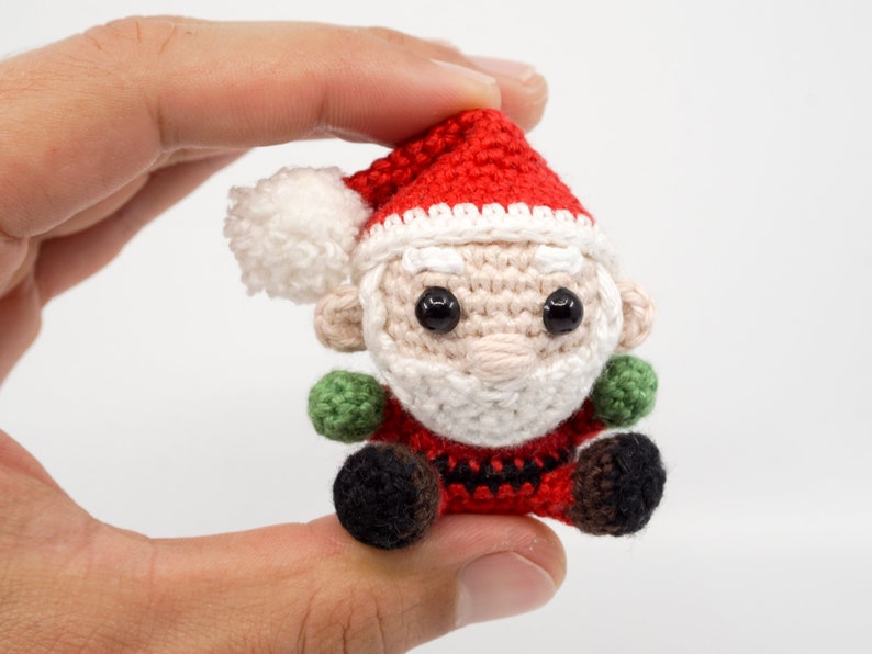 Puede incluir: Un peque&ntilde;o adorno de Pap&aacute; Noel de ganchillo con un gorro rojo, barba blanca y ojos negros. El adorno est&aacute; sentado en una mano.