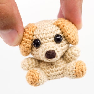 May include: A small crocheted dog toy with brown eyes, a brown nose, and tan ears. The dog is beige with tan accents.