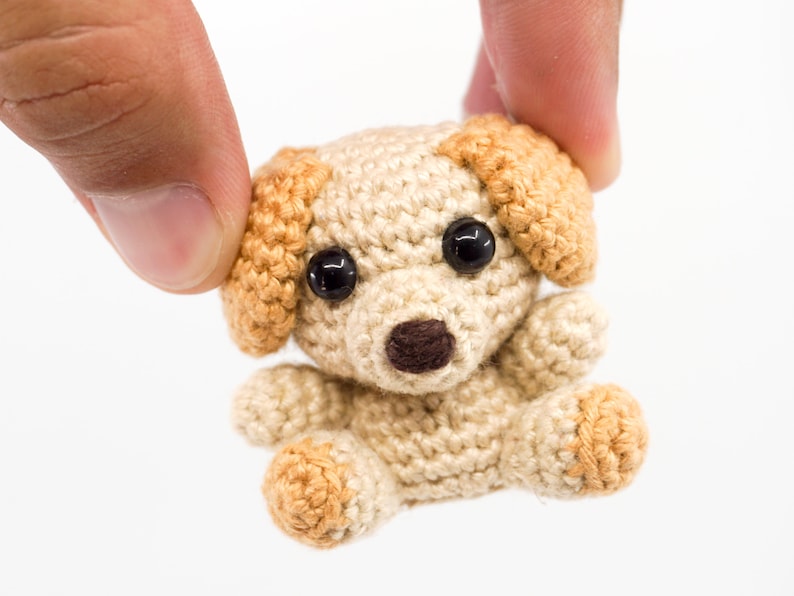 May include: A small crocheted dog toy with brown eyes, a brown nose, and tan ears. The dog is beige with tan accents.