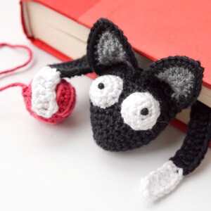 May include: A black crocheted cat bookmark with white eyes and a red yarn ball. The bookmark is hanging off a red book.