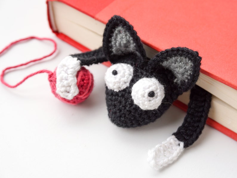 May include: A black crocheted cat bookmark with white eyes and a red yarn ball. The bookmark is hanging off a red book.