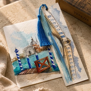 Op de afbeelding: Een rechthoekig aquarel en een bijpassende boekenlegger met een blauw kwastje. Het kunstwerk toont een Venetiaans kanaal met blauw-wit gestreepte meerpalen, een houten steiger en een gebouw met een koepel. De boekenlegger toont een brug.