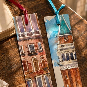 May include: Two rectangular paper bookmarks featuring watercolor illustrations of Italian architecture. The left bookmark shows a multi-story building with red brick tones and a red ribbon. The right bookmark depicts a tall tower with a blue ribbon.