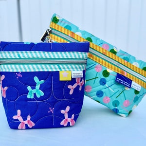 May include: Two quilted zippered pouches with different patterns. One pouch is blue with pink and turquoise balloon dog designs and a label that says "Handmade by Nicole Friend". The other pouch is turquoise with a pattern of pink and blue lollipops and a label that says "Living Life in Bits & Pieces".