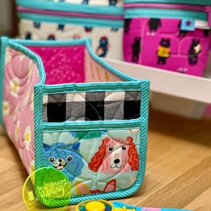 May include: A turquoise and pink fabric storage box with a black and white checked pattern. The box is filled with fabric scraps and has a cute cat and dog design. A yellow rotary cutter and a green circle template with the text "Sea Sassy Stitchery" are in the foreground.