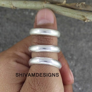 Puede incluir: Tres anillos plateados lisos y pulidos apilados en un dedo. Los anillos tienen un diseño de banda simple y redondeada con un acabado metálico reflectante. El texto SHIVAMDESIGNS está impreso en negro sobre la piel cerca de la base.