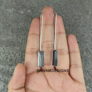 Könnte beinhalten: Ein Paar langer, silberner Ohrringe mit rechteckigen, dunkelgrauen Steinanhängern. Die Ohrringe werden vor einem neutralen Hintergrund präsentiert. Der Text "SHIVAMDESIGNS" ist sichtbar.