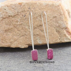 Könnte beinhalten: Silberne Ohrringe mit einem langen, dünnen Drahtdesign. Jeder Ohrring hat einen rechteckigen, grob geschnittenen, rosa Edelstein. Die Ohrringe sind vor einem strukturierten, hellbraunen Steinhintergrund platziert. Der Text "SHIVAMDESIGNS" ist sichtbar.